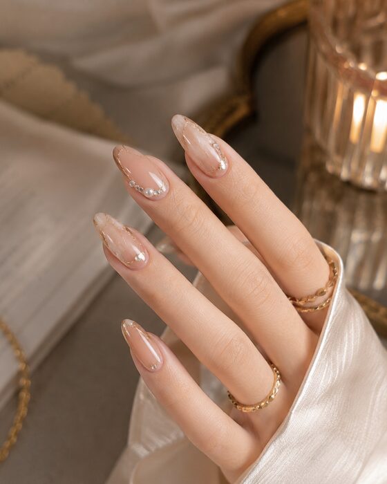 Sheer nude almond nails with delicate crystal details and gold rings